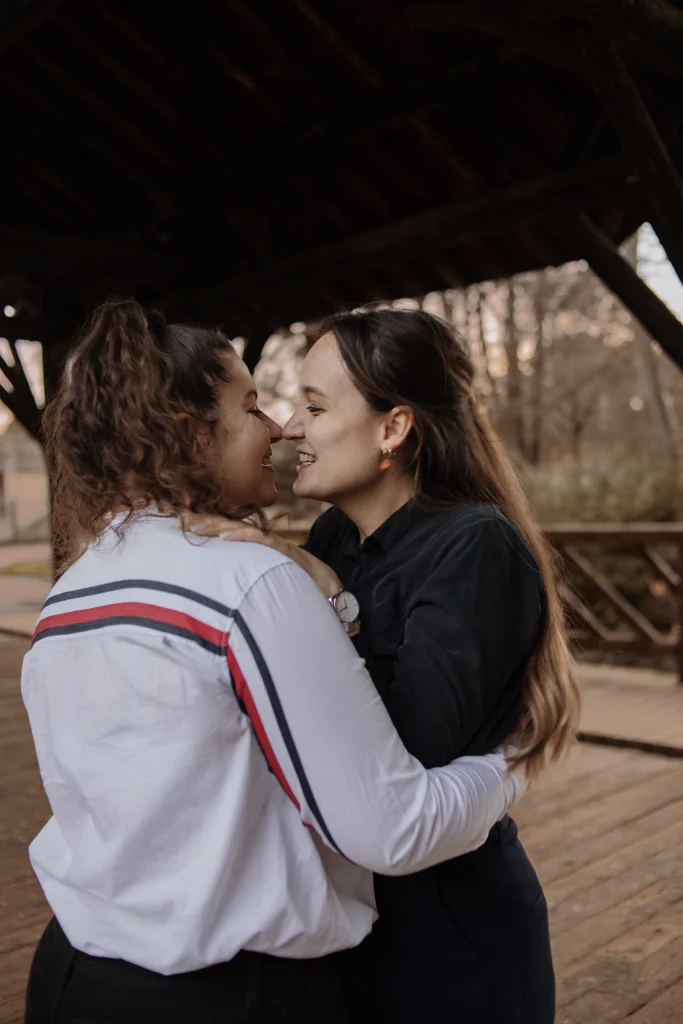 Deux femmes enlacées, échangeant un regard complice sous une charpente en bois, en extérieur.