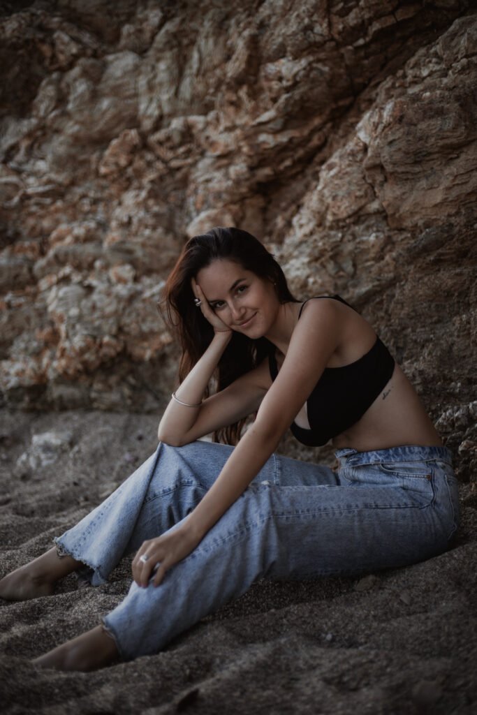 Jeune femme souriante, assise en jean et top noir contre une paroi rocheuse en bord de mer.