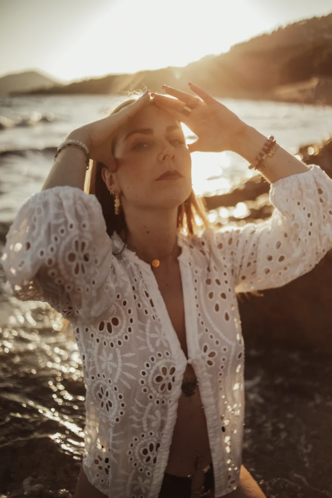 Femme en robe blanche ajourée, bras levés face au soleil couchant.