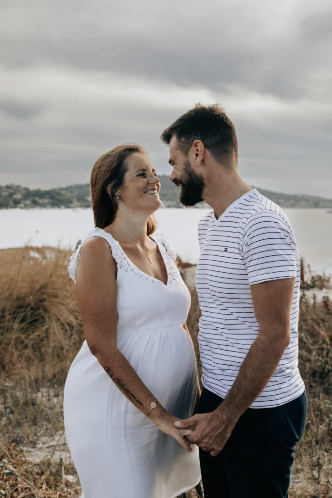 Un couple se tenant debout, l'un en face de l'autre, main dans la main sur la plage. Ils se regardent avec tendresse et ris.