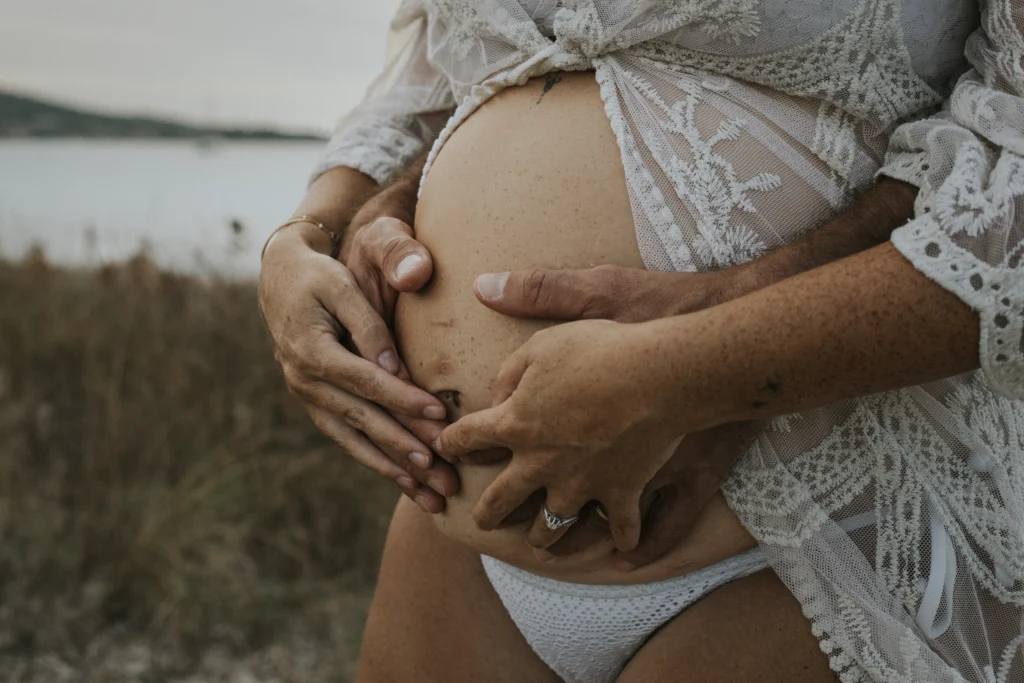 Ventre rond d'une femme enceinte sur lequel sont posées les mains entrelacées des futurs parents. En arrière plan, on distingue la végétation et la mer, un endroit parfait pour une séance photo grossesse.