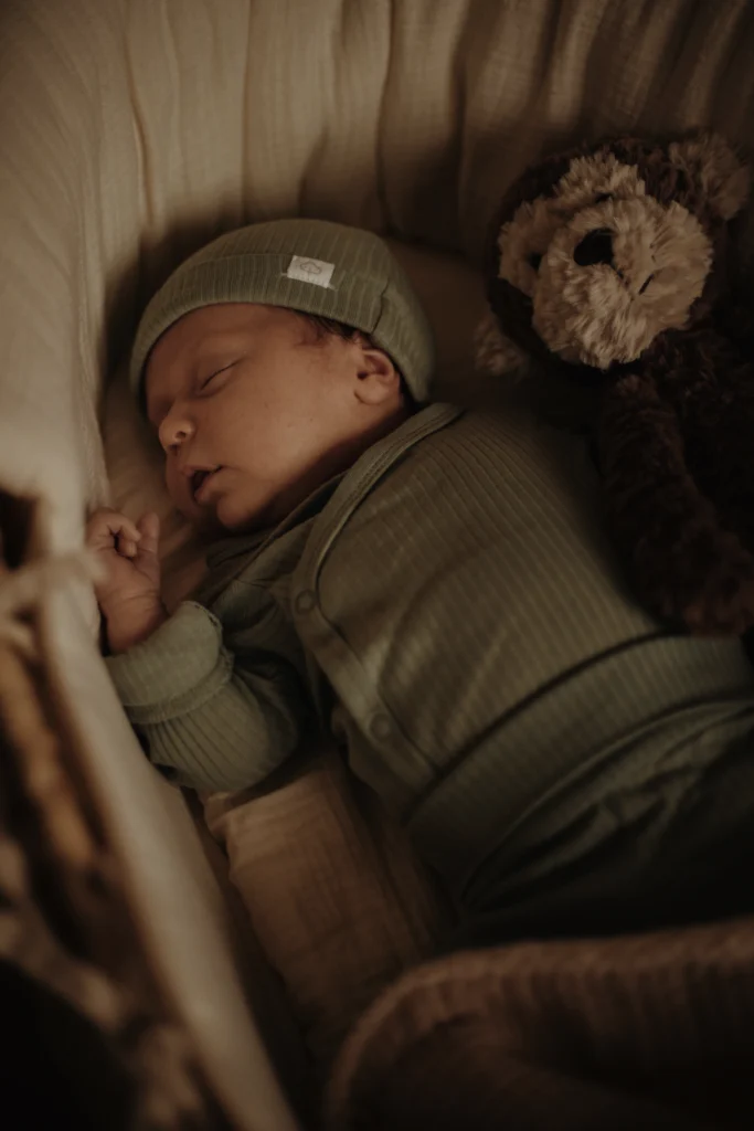 Un nourrisson vêtu d’un ensemble vert et d’un bonnet assorti dort paisiblement dans un couffin, accompagné de son doudou en peluche.