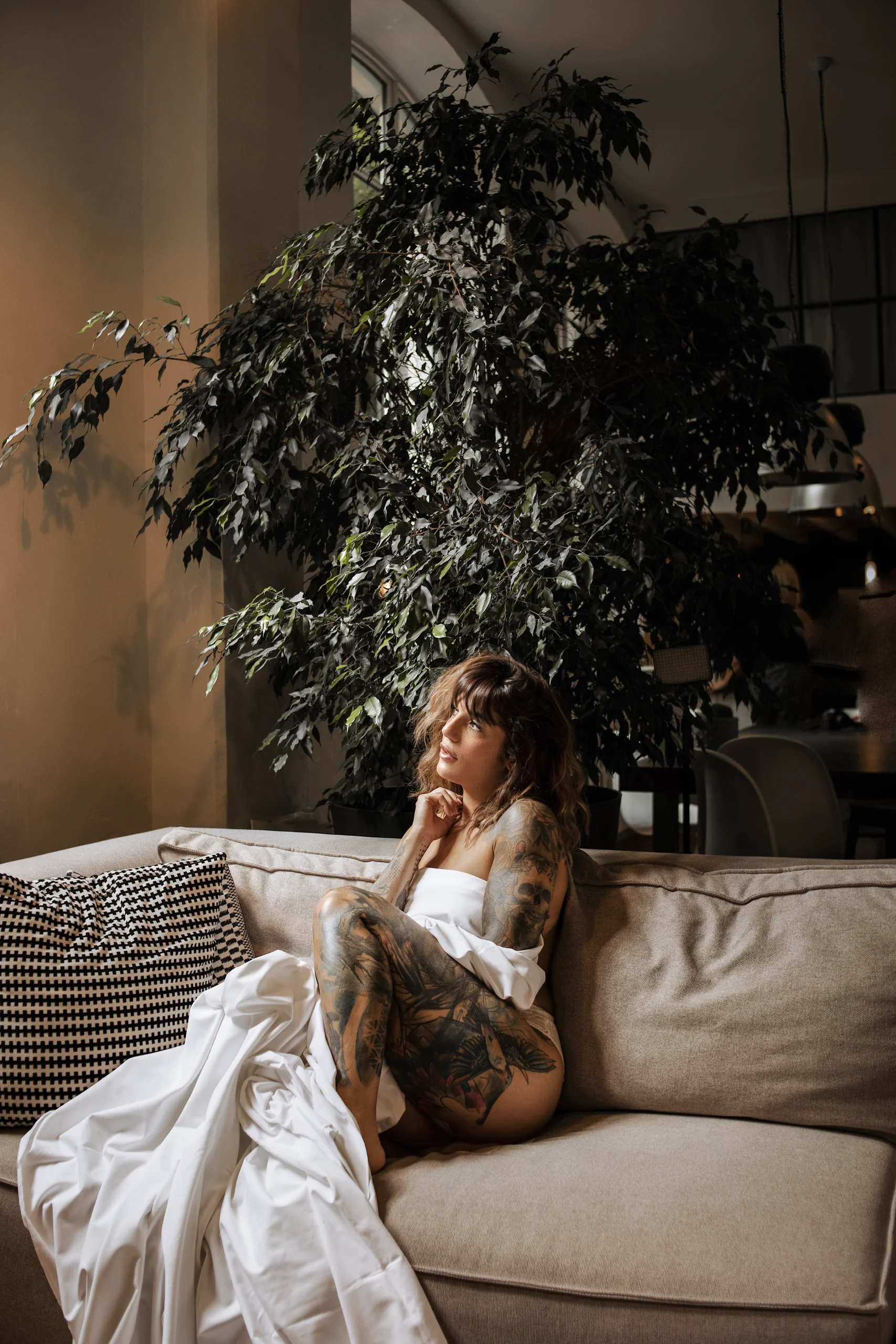 Une jeune femme, couverte d'un long tissu blanc, est assise sur un canapé, devant une grande fenêtre, dans un appartement moderne.