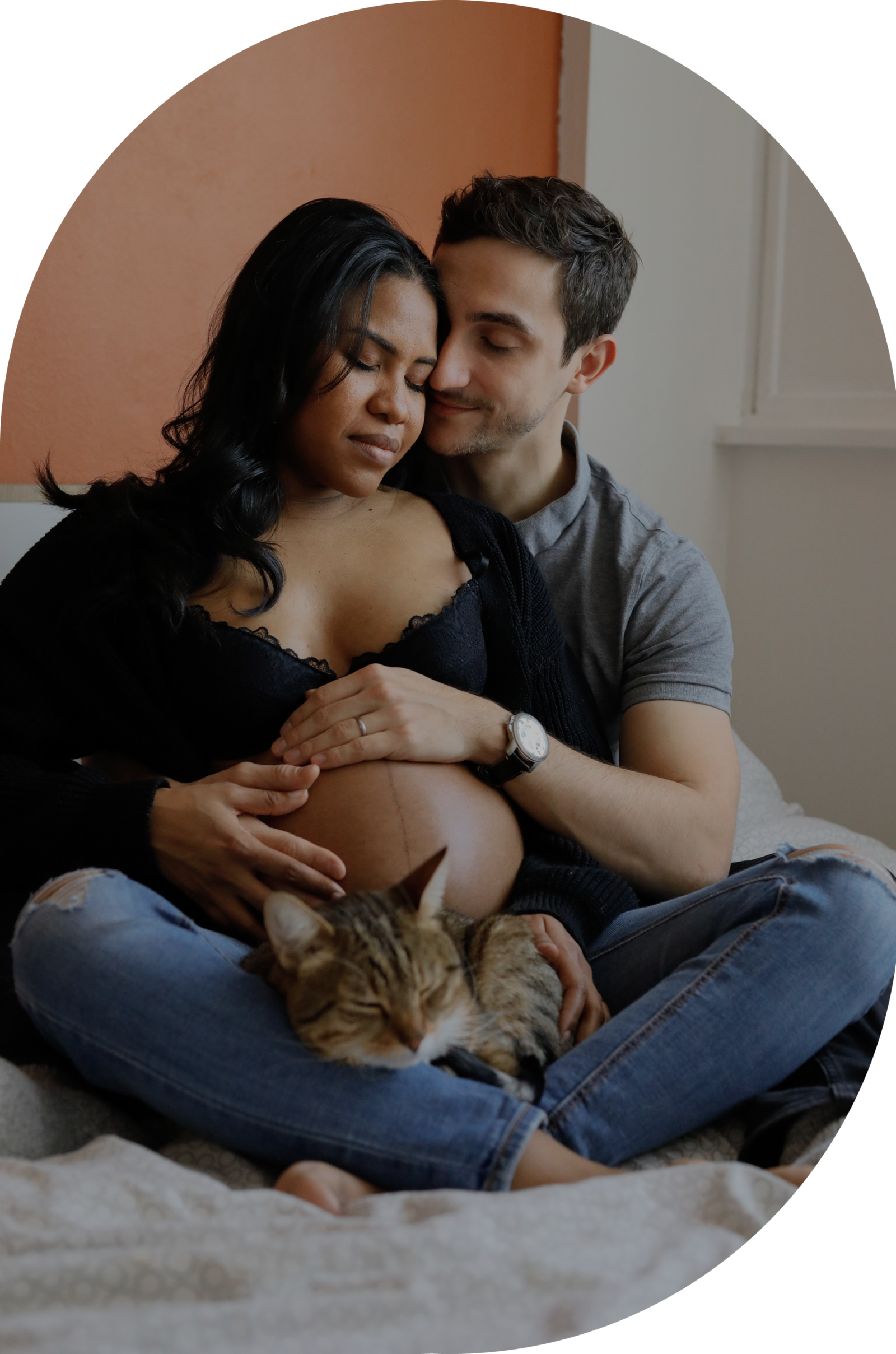 Un couple en attente d'un enfant, assis l'un contre l'autre sur un lit dans une chambre cosy, accompagné de leur chat.