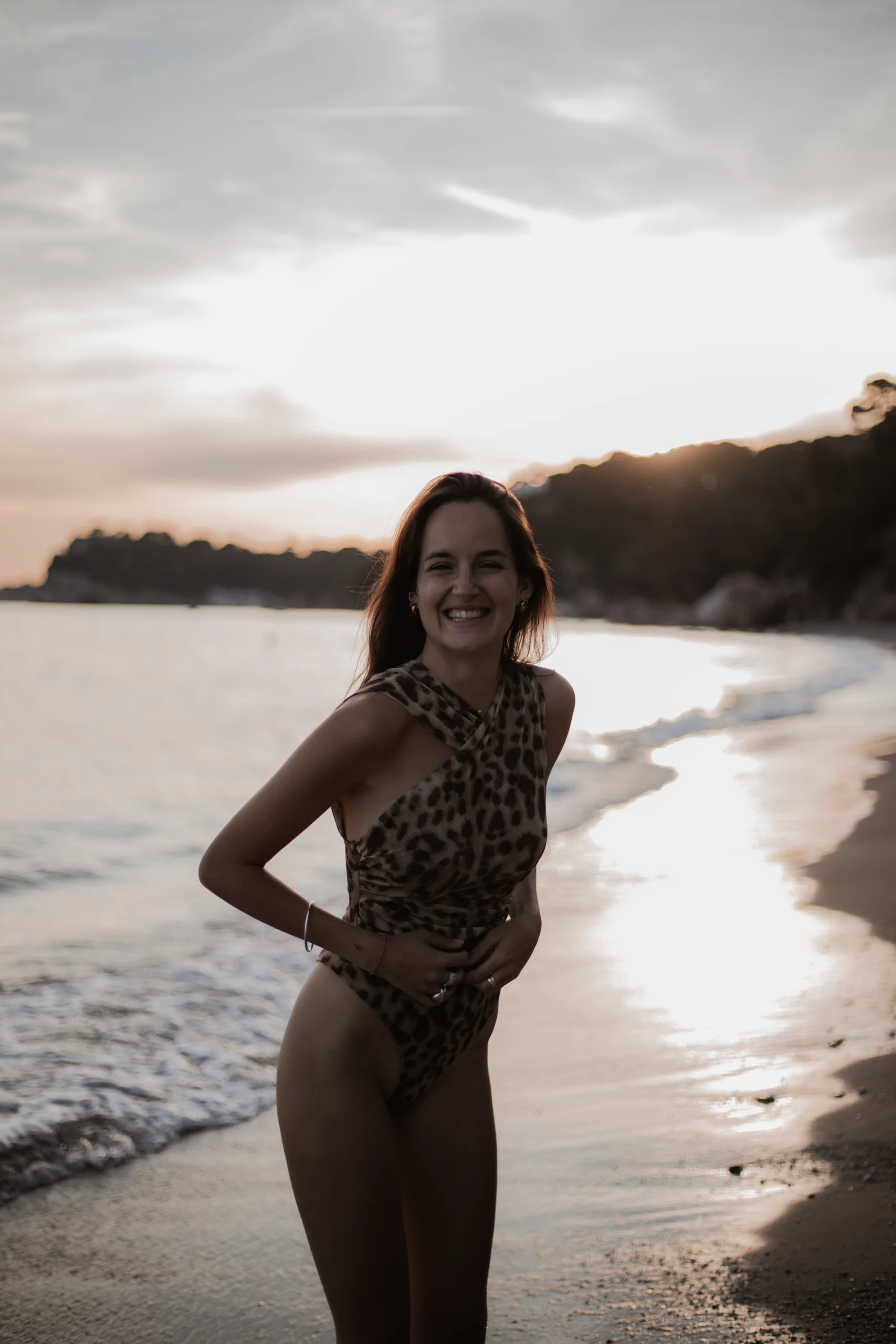 Une femme se tenant debout sur la plage en maillot et riant devant l'objectif. Elle est elle même car elle a une bonne relation client photographe.