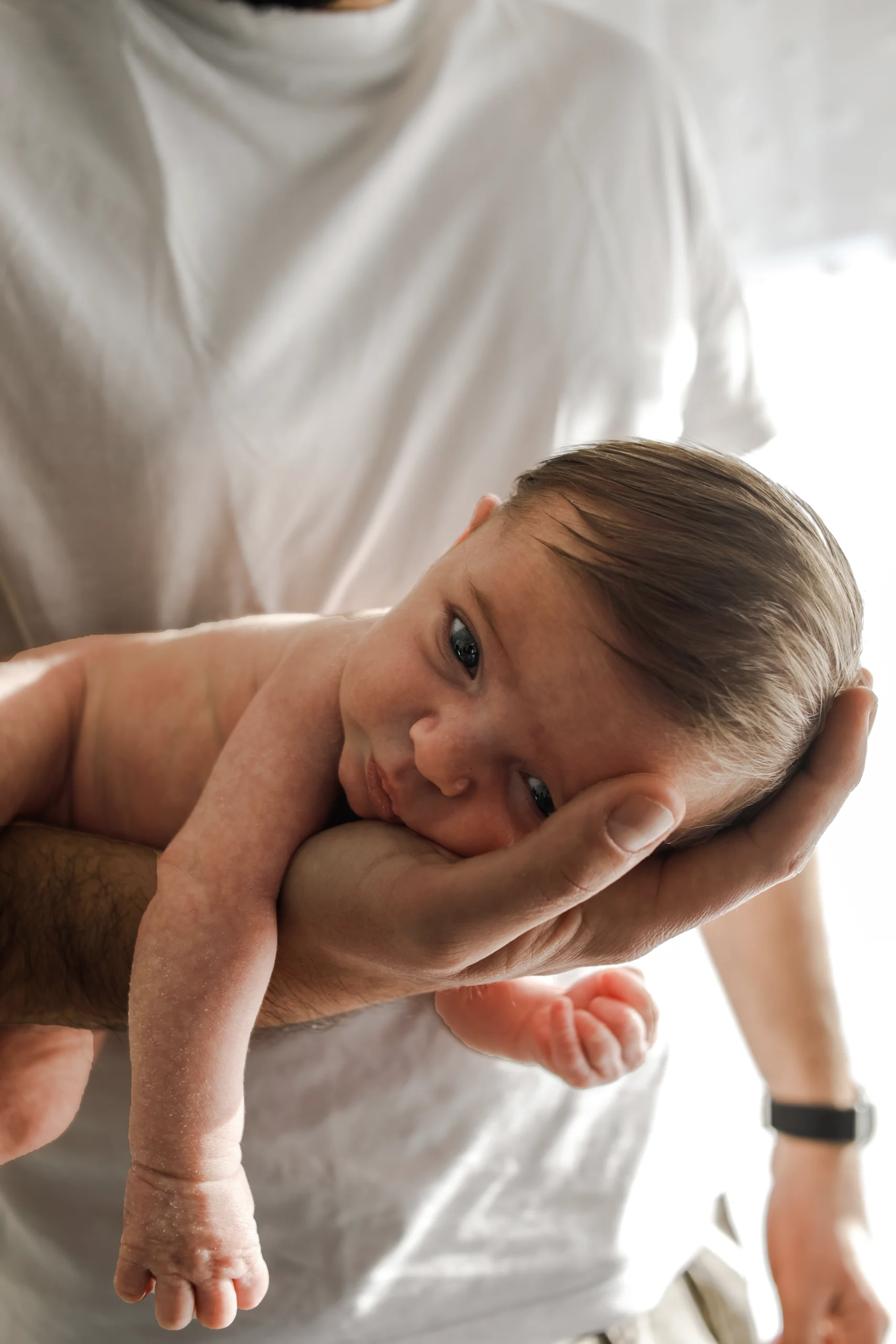 Un nouveau né, la tête dans la paume de la main sur son père, est allongé sur le ventre, sur le bras de son père lors d'une séance photo naissance lifestyle.