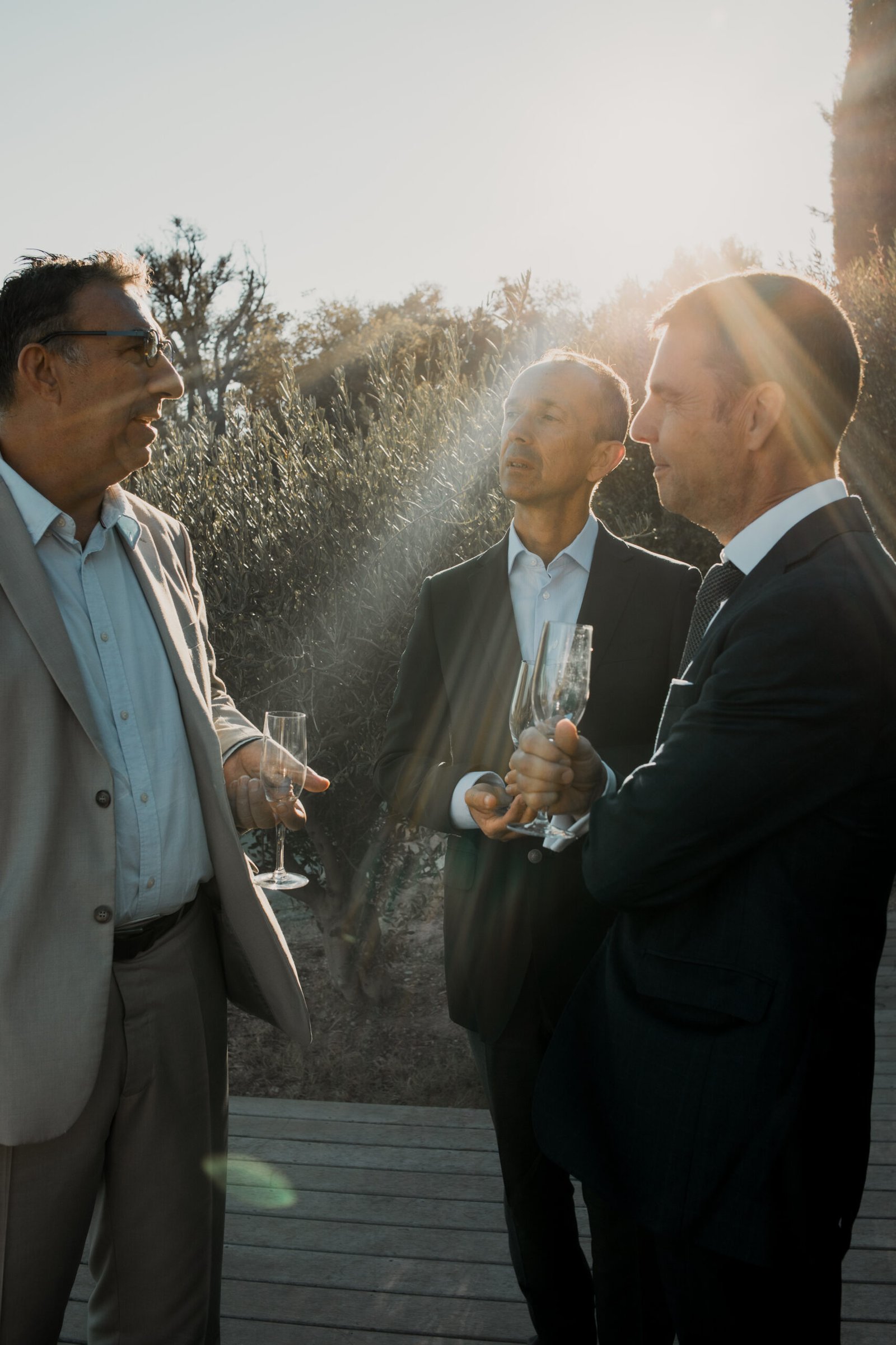 Trio d'hommes en costume lors d'un mariage en Provence. Ils se tiennent devant le soleil couchant, un verre à la main durant les photos de groupes.