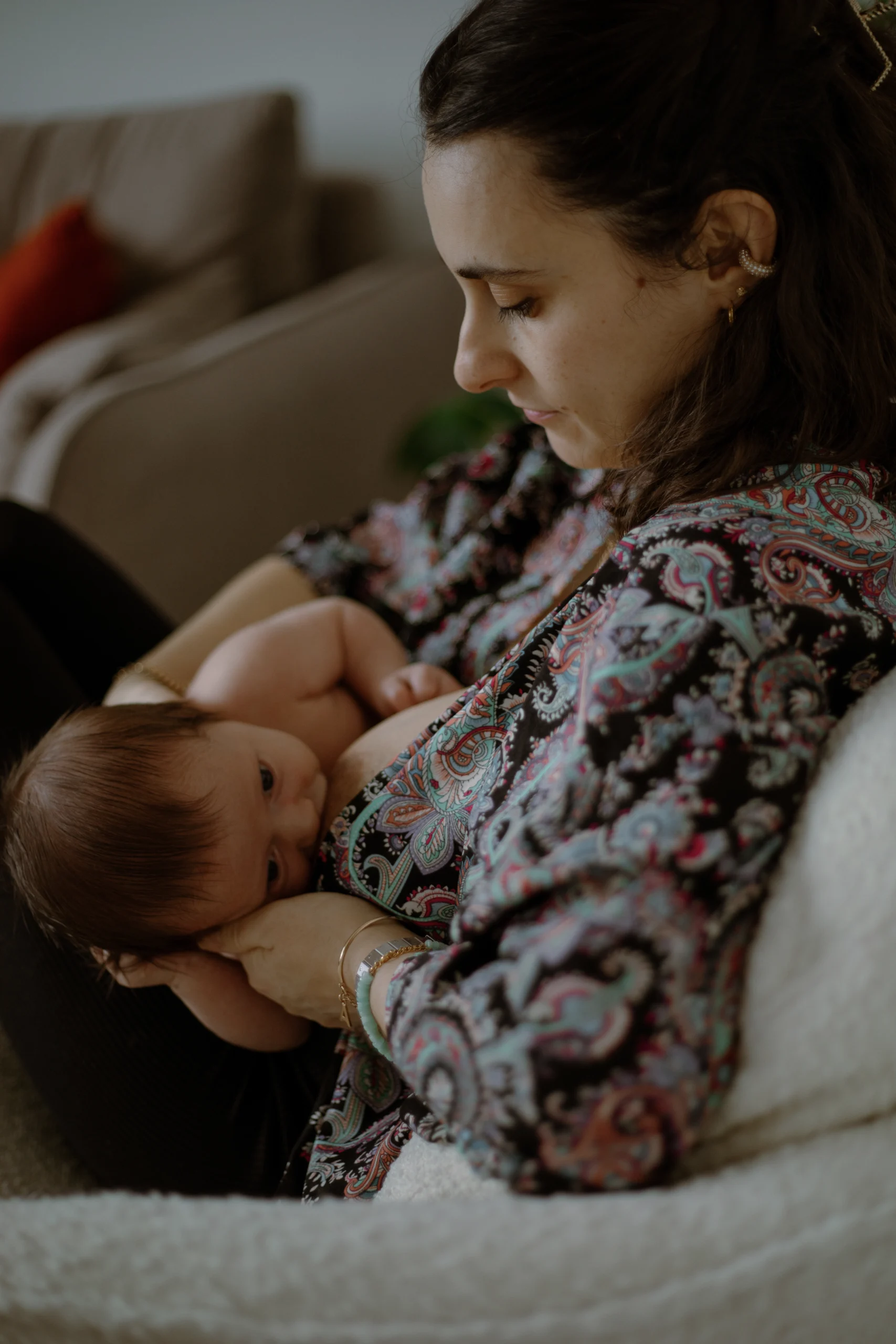 Une mère, allaitant son enfant, assisse sur un canapé. Un moment doux et intime lors d'une séance photo naissance lifestyle.
