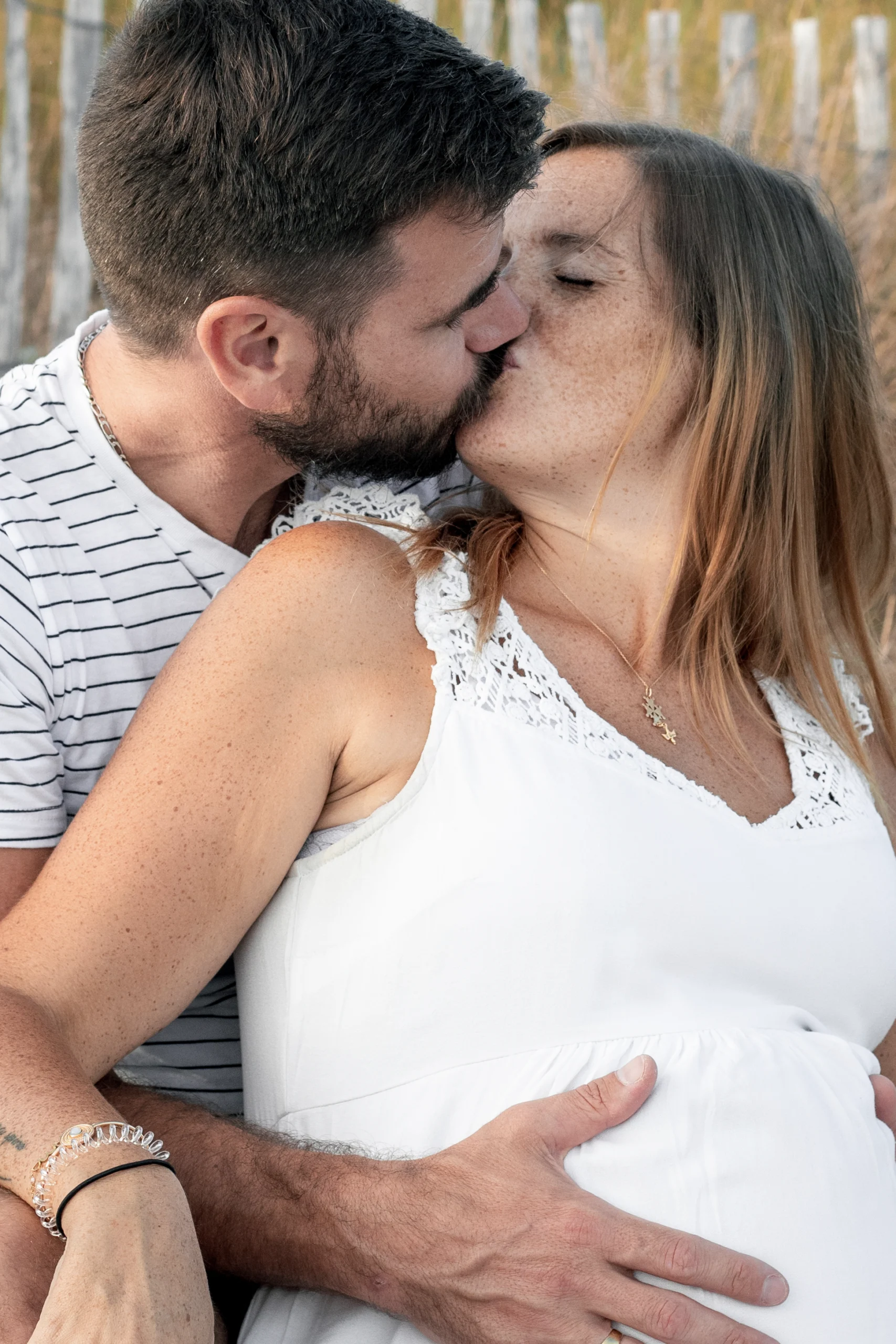 Un couple attendant un bébé, s'embrasse tendrement assis sur le sable. Ils sont à l'aise et ne ressentent pas de gêne car ils ont construit une vraie relation client photographe.