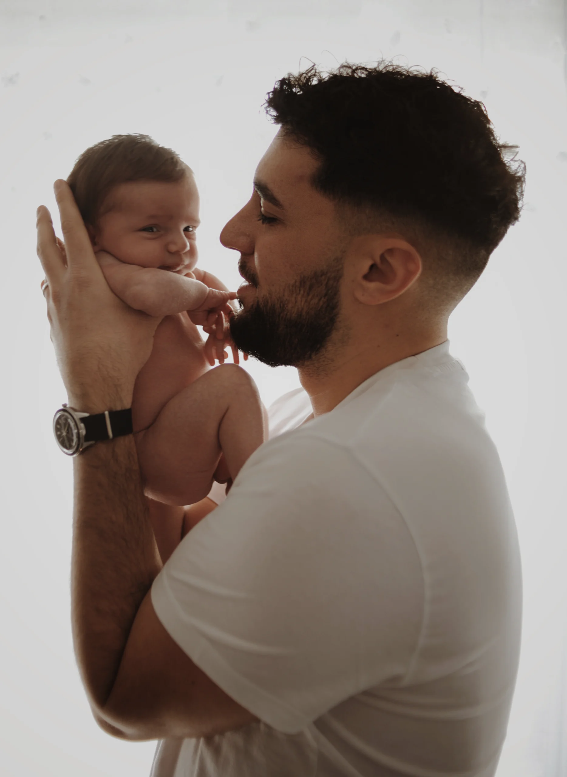 Un père, portant son bébé recroquevillé près de visage et le regardant avec amour lors d'une séance photo naissance lifestyle.