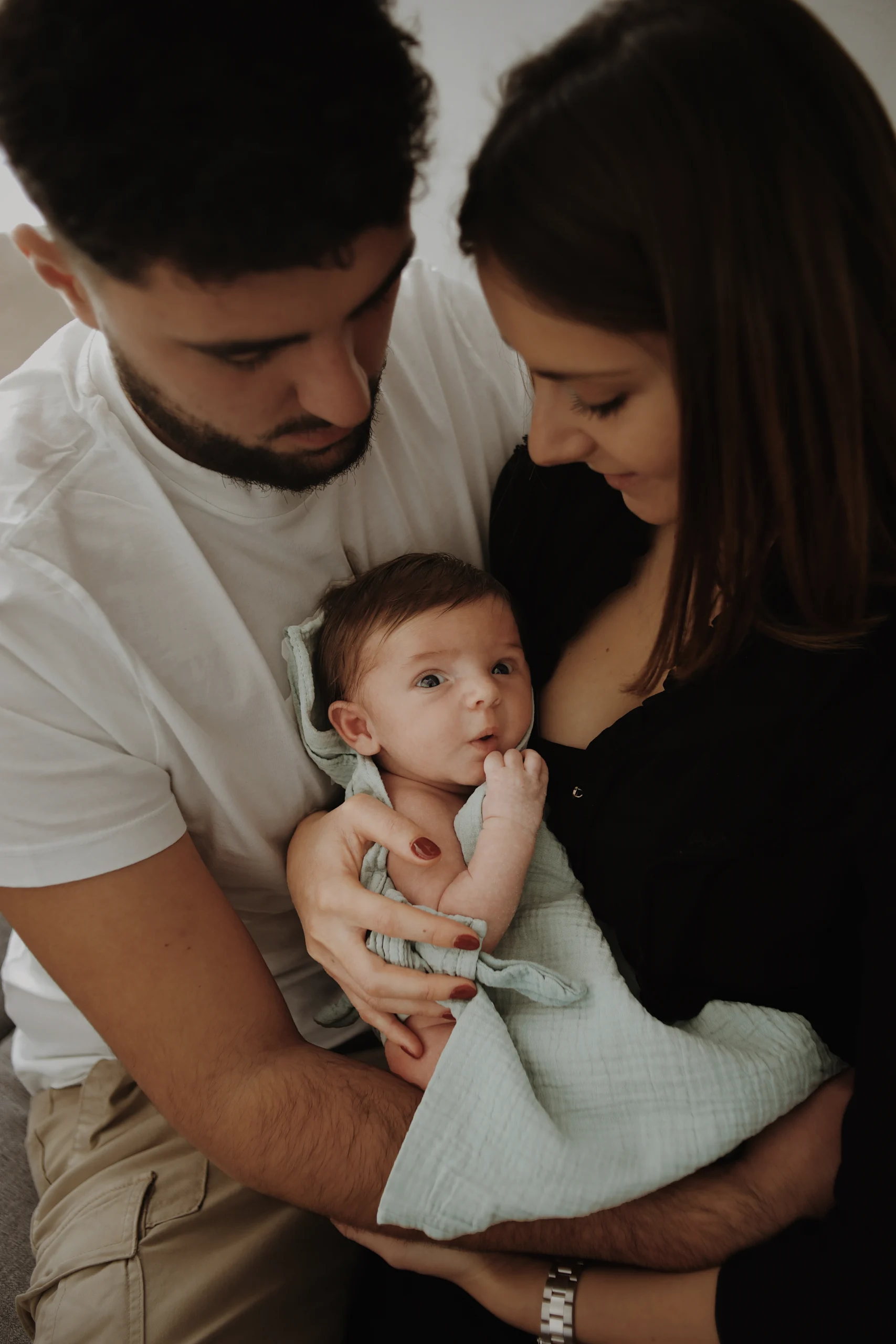 Un couple de jeune parents tiennent dans leur bras leur bébé nouveau né, enveloppé d'une couverture vert d'eau. Une séance photo naissance lifestyle.
