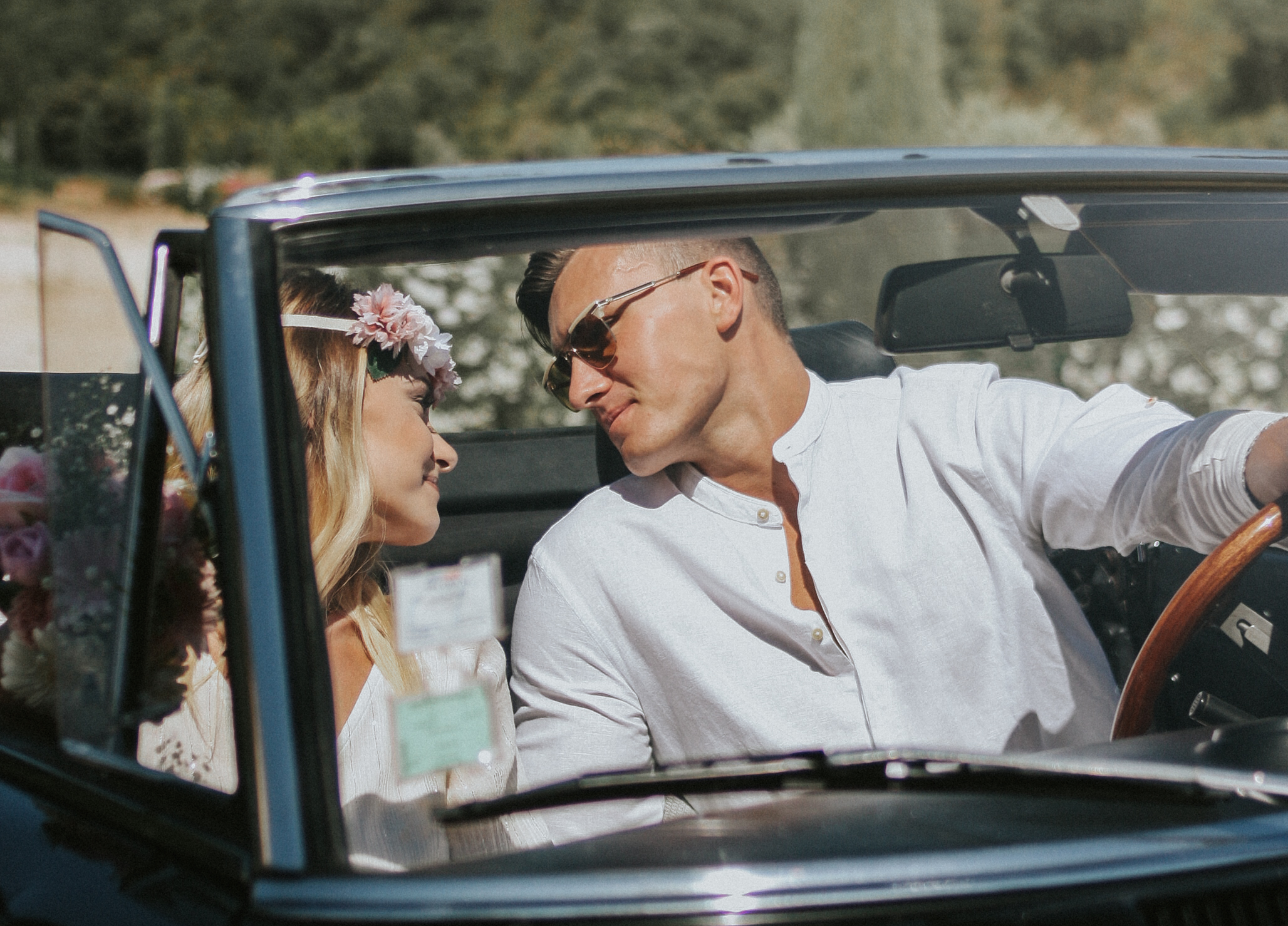 Un couple, au volant d'un voiture vintage décapotable, se regarde amoureusement. Ils portent leurs tenues de mariages. La femme porte une couronne de fleurs.