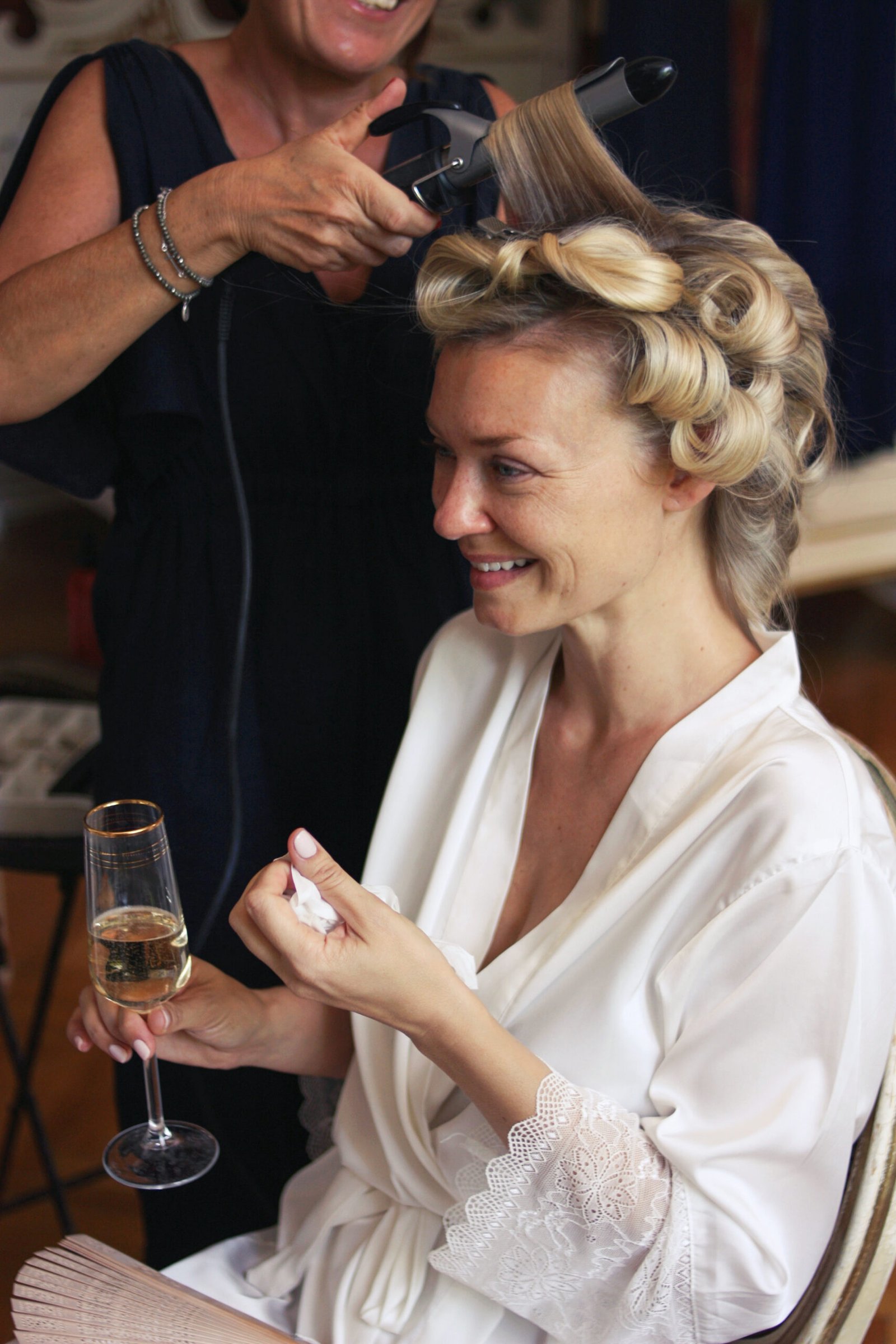 Une mariée tenant une coupe de champagne à la main est en train de se faire coiffer pour son mariage. La lumière est naturelle, la femme est placée devant une grande fenêtre.