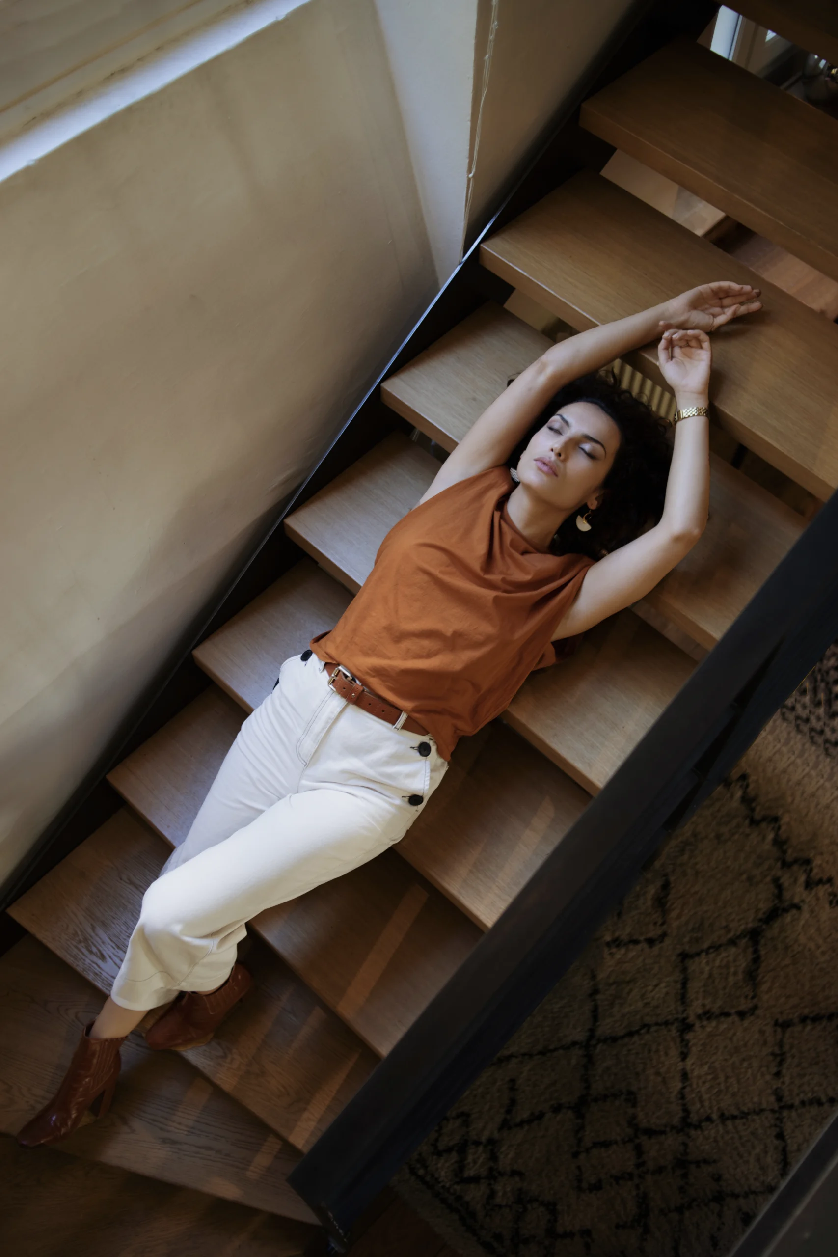 Une jeune femme vêtue d'un pantalon blanc et d'un t-shirt de couleur ocre est allongée dans des escalier modernes, dans un intérieur élégant. Une séance photo confiance en soi en intérieur dans le Var.