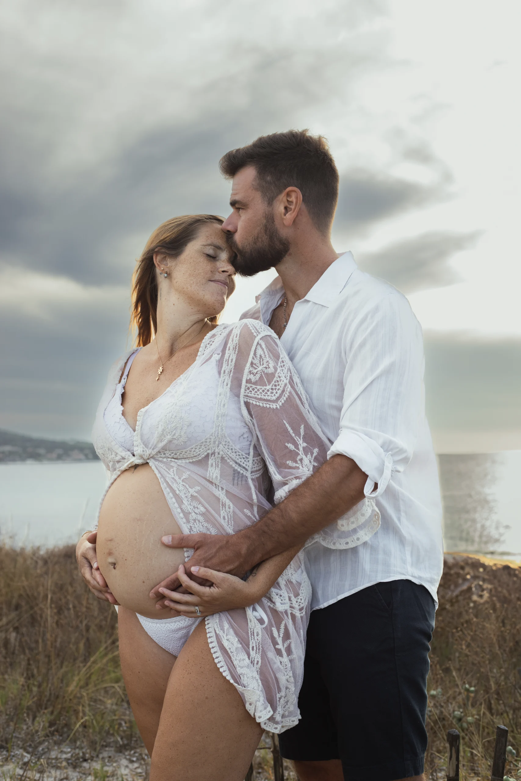 Un couple en bord de mer. L'homme est placé debout derrière sa femme et pose un bisous sur son front. Ils ont les mains posés sur le ventre rond.