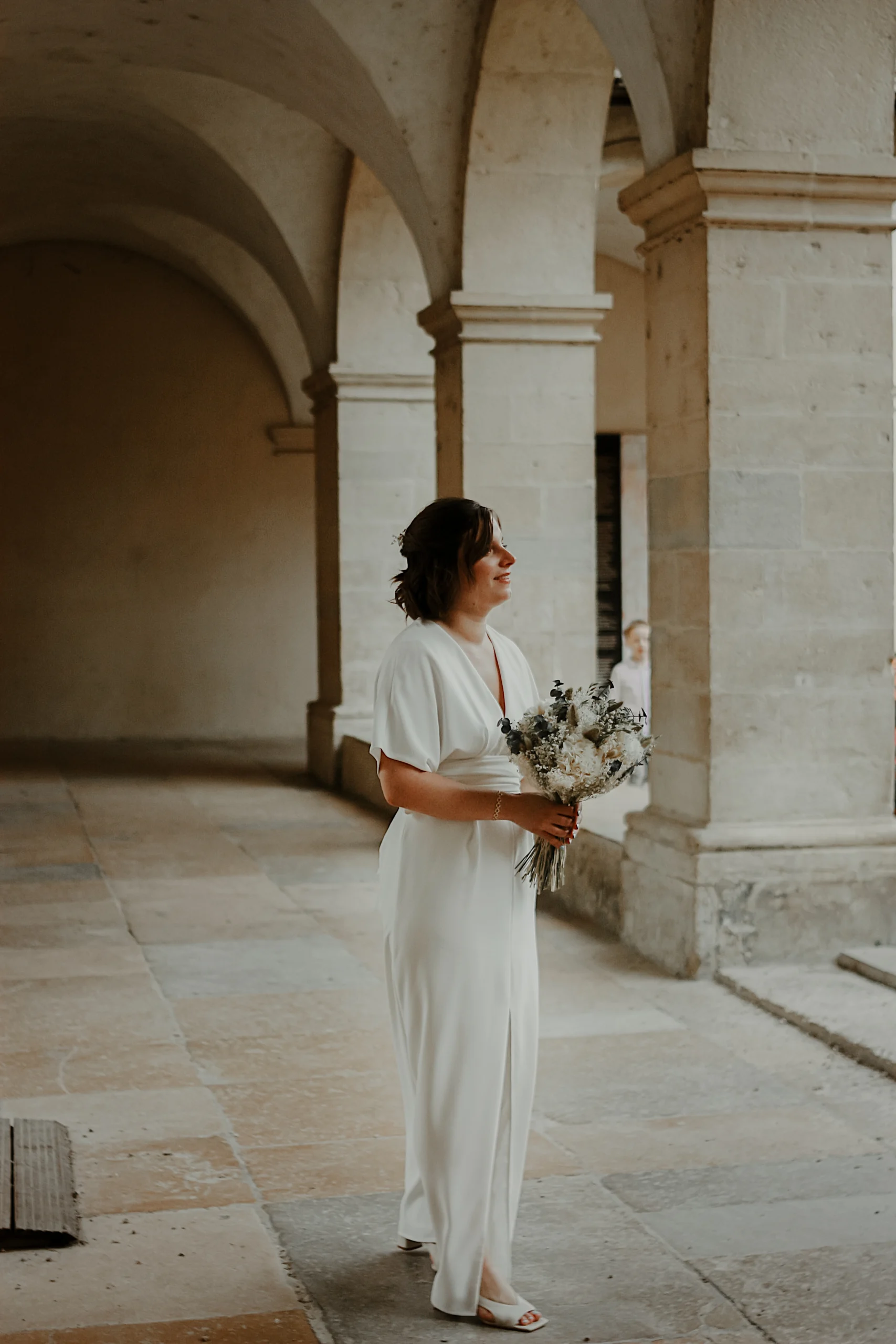 Une mariée en robe blanche, sous les voute d'un bâtiment ancien, tenant son bouquet bohème dans les mains.