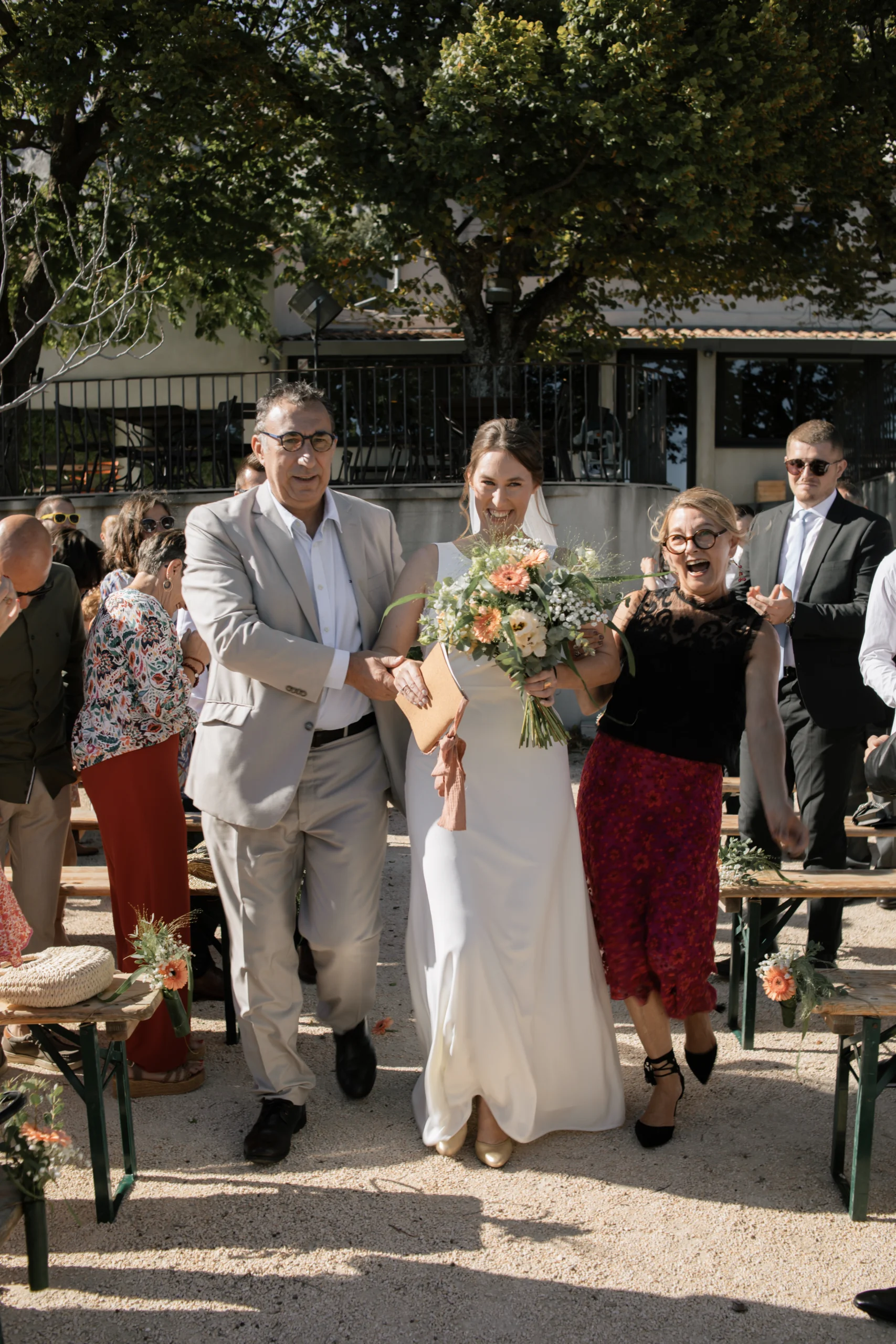 Une mariée, descendant l'allée de sa cérémonie laique lors de son mariage en Provence. Elle est accompagnée de ses parents. C'est une photographie de mariage naturelle et pleine de vie.