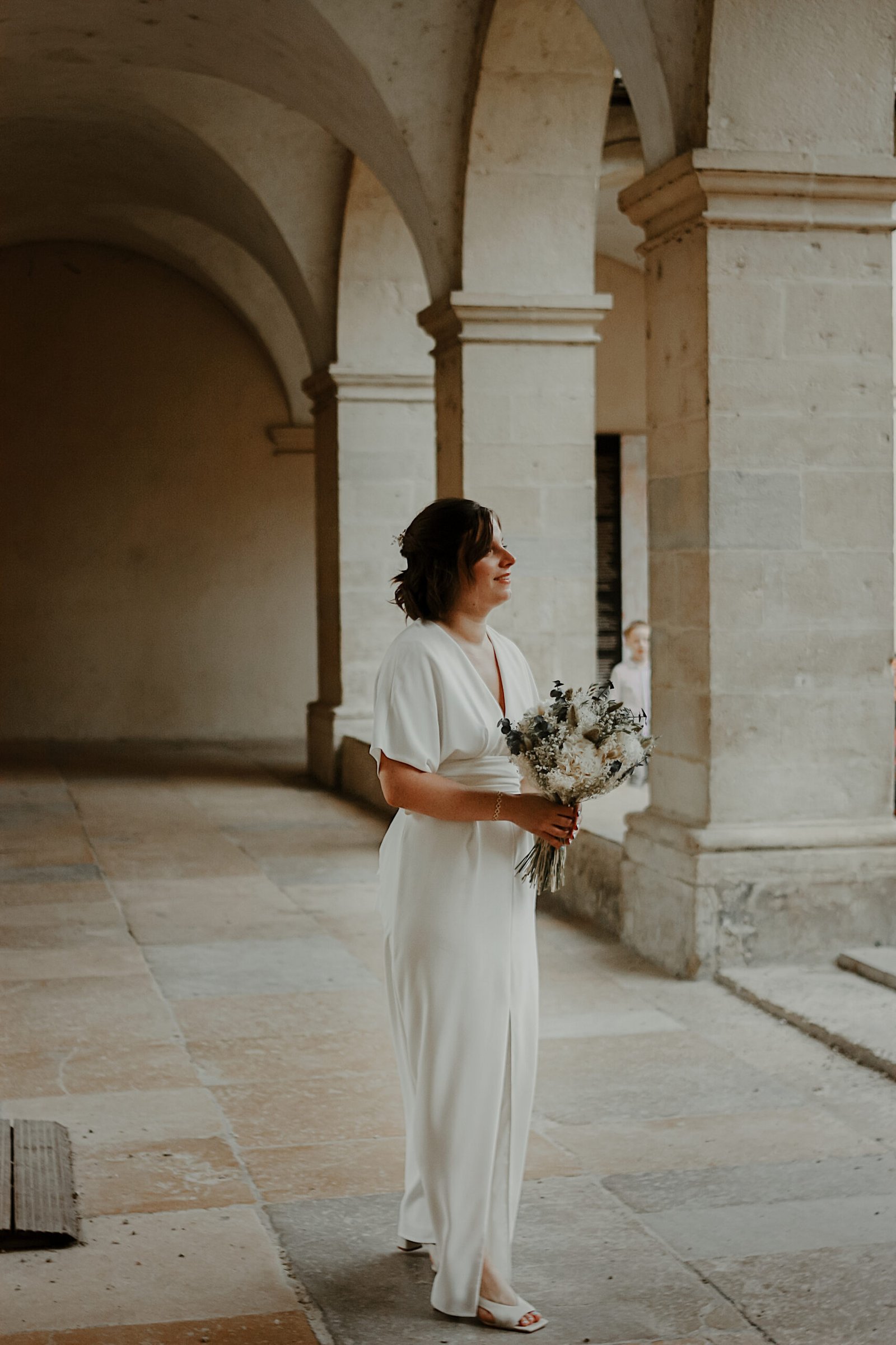 Une mariée, sous les arche d'un bâtiment, son bouquet à la fin. Photo prise par une photographe de mariage à Toulon.