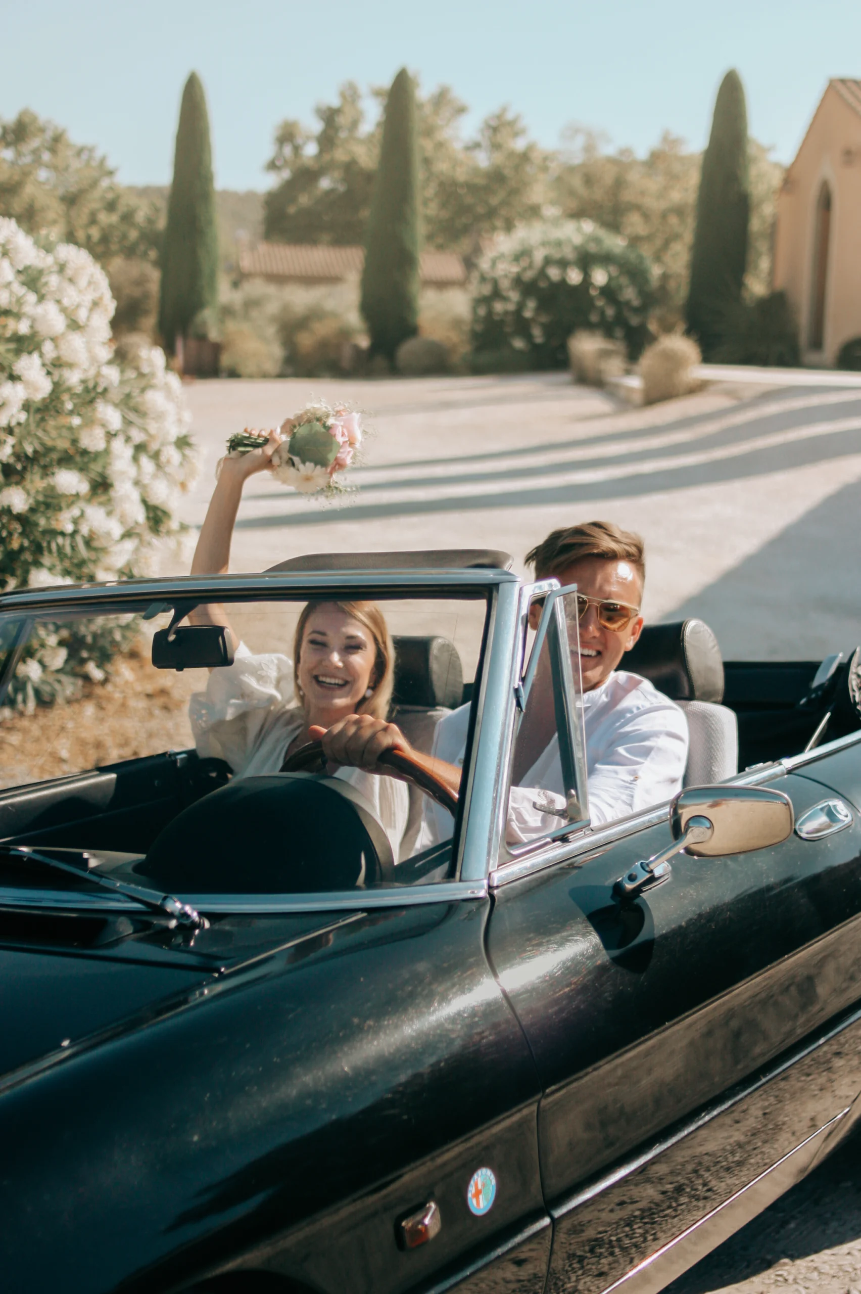 Une photo de mariage dans un style documentaire, lors d'un mariage en Provence. Les mariés sont dans une voiture noire décapotable et rient.