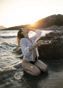 Une jeune femme agenouillée au bord de l'eau porte un gilet en maille blanc et un maillot de bain noir.