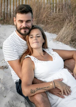 Un couple assis l'un contre l'autre sur la plage, caressant le ventre rond de la femme lors d'une séance grossesse à Hyères. Ils portent des tenues associées.